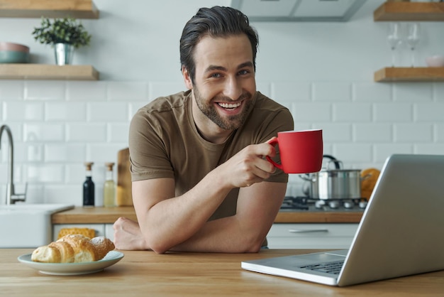 Hombre alegre disfrutando del café de la mañana mientras está sentado en la isla de la cocina