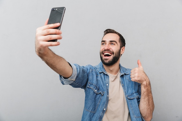 Hombre alegre emocionado con camisa aislada sobre pared gris, tomando un selfie