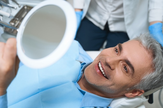 Hombre alegre está acostado en una silla y mirando en el espejo mientras está encantado con el trabajo del dentista