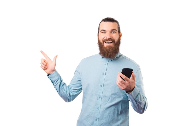 El hombre alegre está apuntando sobre el fondo blanco mientras sostiene su teléfono.