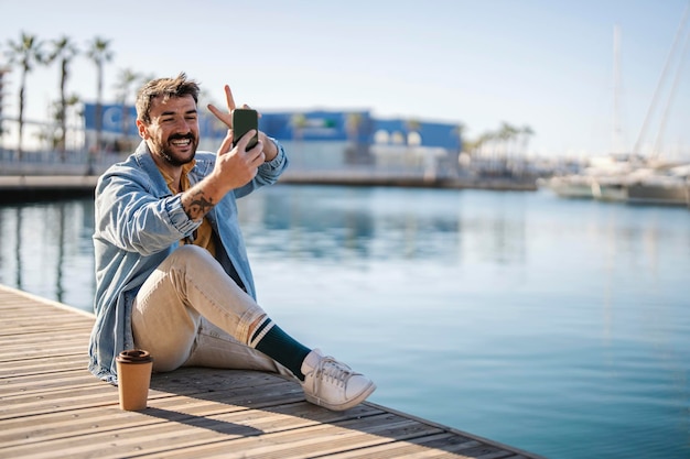 Un hombre alegre está sentado en un muelle y tomando selfies