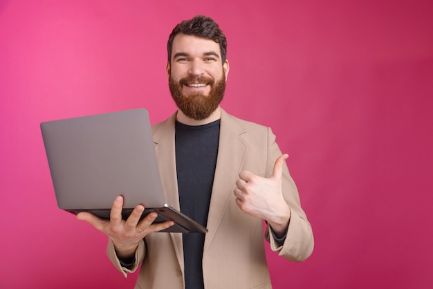 El hombre alegre está sosteniendo su computadora portátil y está mostrando como gesto.