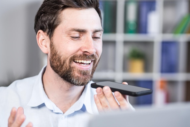 Hombre alegre y feliz graba mensajes de audio empresario independiente en la oficina utiliza la aplicación en el teléfono inteligente