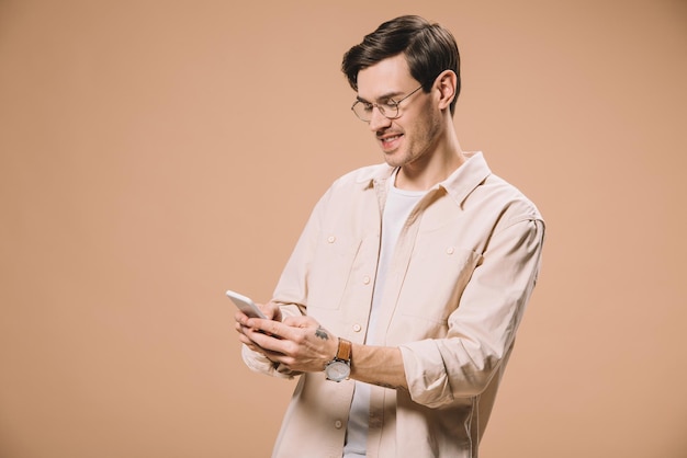 Hombre alegre con gafas mirando el teléfono inteligente aislado en beige