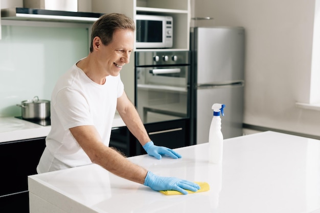 Hombre alegre con guantes de goma sosteniendo un trapo mientras limpia la mesa de la cocina