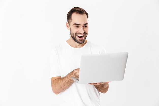 Hombre alegre guapo vestido con camiseta en blanco que se encuentran aisladas sobre la pared blanca, usando la computadora portátil