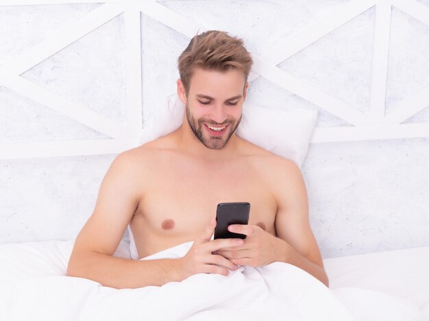 Hombre alegre leyendo un mensaje por teléfono en un dormitorio blanco