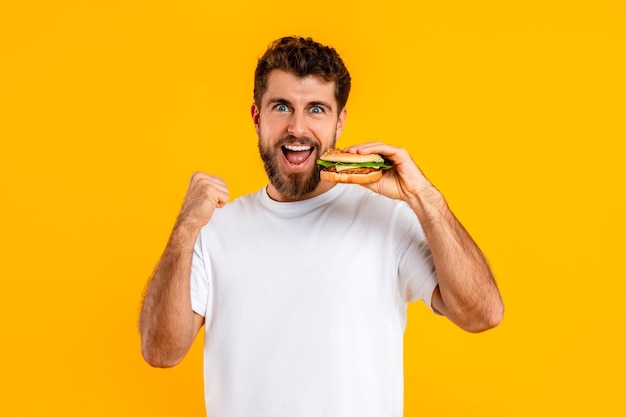 Foto hombre alegre mordiendo una hamburguesa con queso y haciendo gestos de sí sobre un fondo amarillo