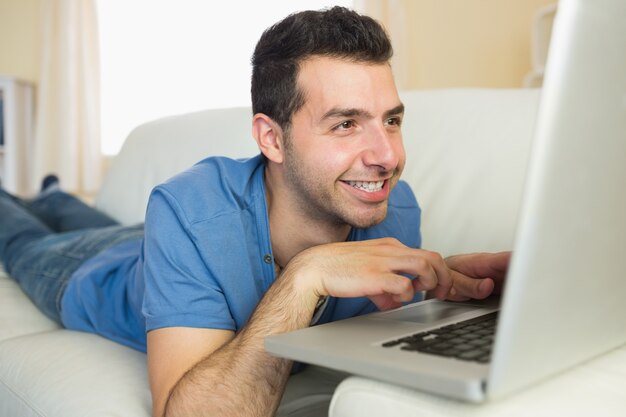 Hombre alegre ocasional que se sienta en el sofá usando y mirando la computadora portátil