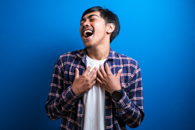 El hombre alegre positivo cerrando los ojos con fuerza no puede dejar de reírse de una buena broma. Apuesto joven de apariencia asiática con cabello negro con camisa de franela se encuentra sobre fondo azul en Studio