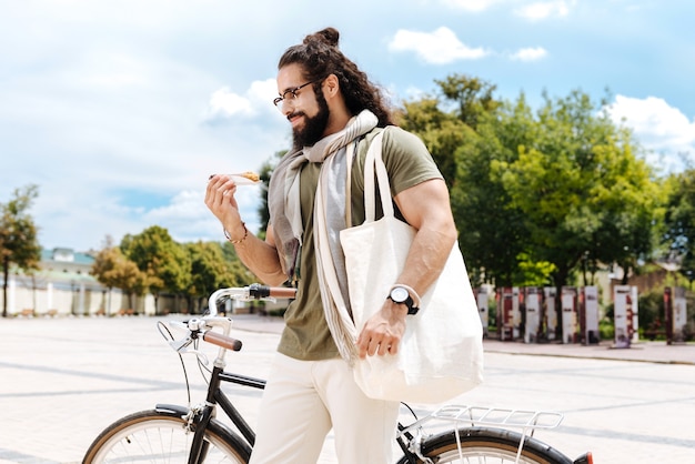 Hombre alegre positivo de pie cerca de su bicicleta mientras come comida de la calle