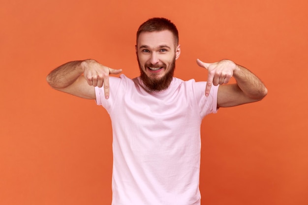 Un hombre alegre y satisfecho señalando con el dedo hacia abajo prestando atención al blogger del área de publicidad que recomienda suscribirse usando una camiseta rosa Foto de estudio interior aislada en un fondo naranja