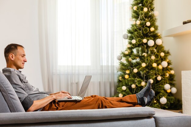 Hombre alegre sentado en el sofá y usando la computadora portátil cerca del árbol de Navidad en casa