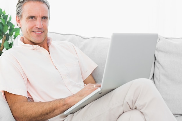 Hombre alegre en su sofá usando la computadora portátil mirando la cámara en casa en la sala de estar