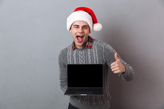 Hombre alegre en suéter y sombrero de navidad que muestra la pantalla de la computadora portátil en blanco y el pulgar hacia arriba