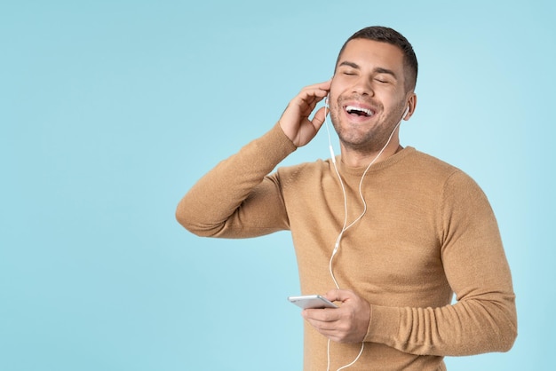 Hombre alegre con teléfono y auriculares disfrutando de la música con los ojos cerrados aislado sobre fondo azul.