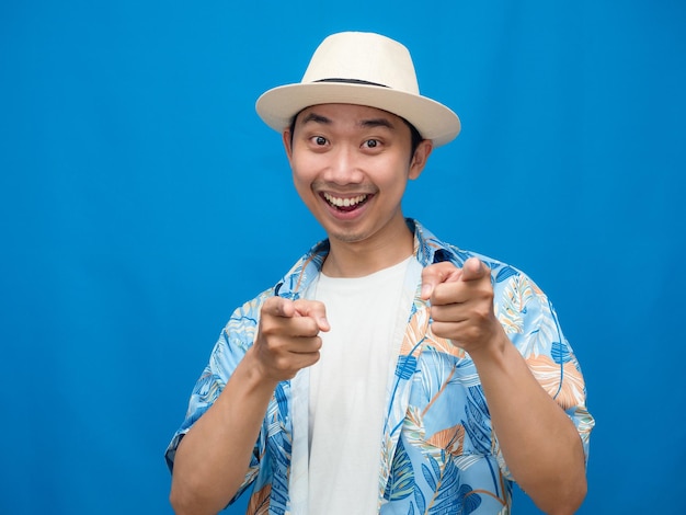 Un hombre alegre usa un sombrero, una camisa de playa, te señala con el dedo el fondo azul.