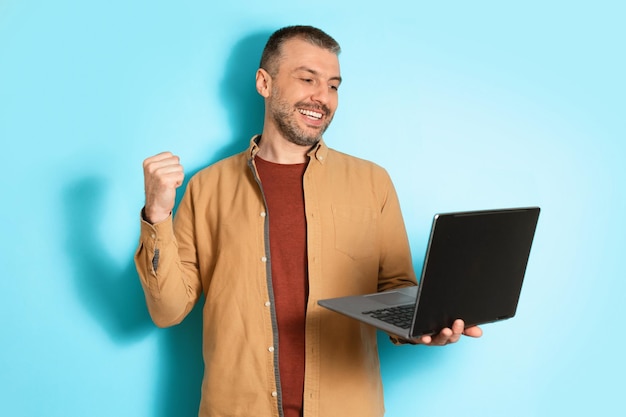 Hombre alegre usando laptop sacudiendo puños sobre fondo azul.