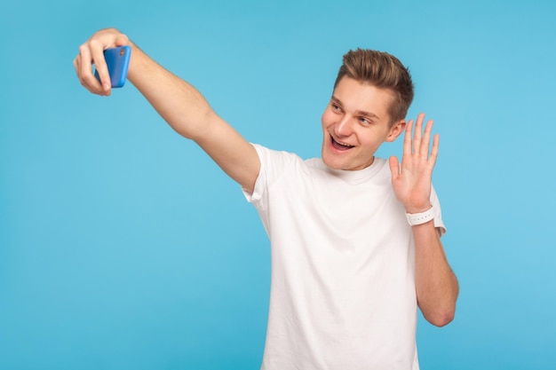 Un hombre amable y feliz con una camiseta blanca informal saludando mientras habla por videollamada haciendo selfie usando un teléfono móvil en línea chateando en un estudio interior aislado en un fondo azul
