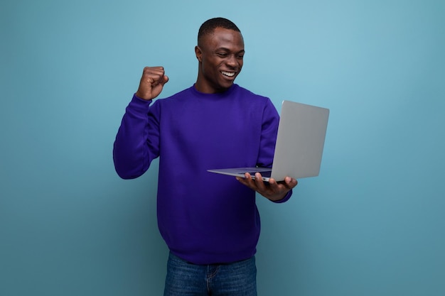 Un hombre americano de piel oscura vestido con un suéter azul trabaja remotamente con una computadora portátil