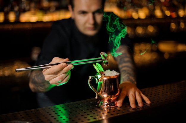 Hombre añadiendo a un cóctel en el vaso de cobre con una canela ahumada aromática naranja seca con pinzas en la barra del bar