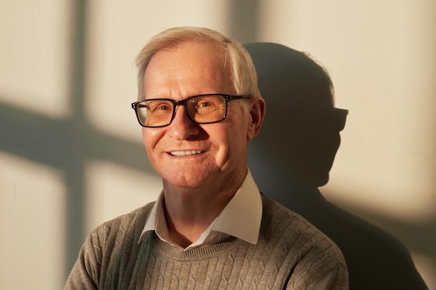 Hombre anciano alegre con anteojos y jersey casual de pie junto a la pared