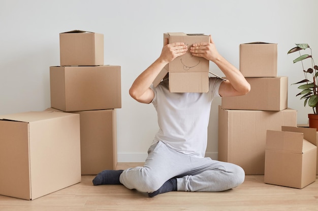 Un hombre anónimo desconocido con camiseta blanca sentado en el suelo cerca de cajas de cartón, lleva una caja de cartón con la cara dibujada y cubre los ojos con las palmas, divirtiéndose durante la mudanza a un nuevo piso.