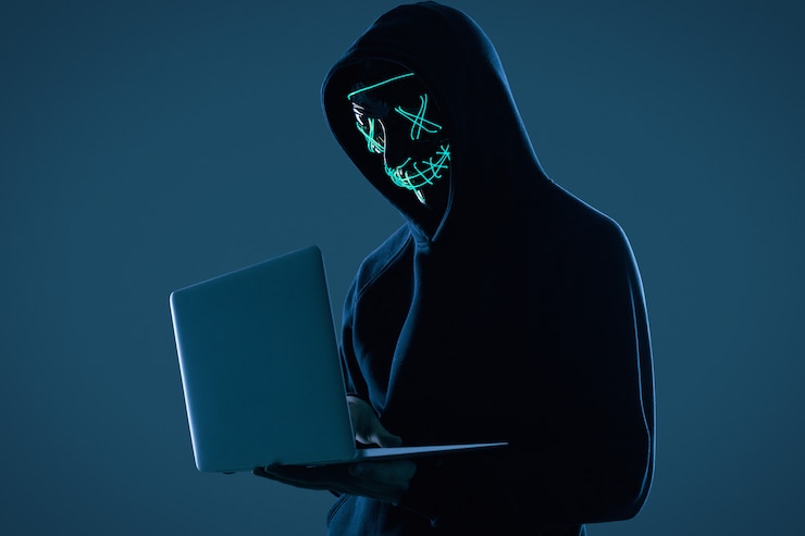 Foto hombre anónimo con una sudadera negra y una máscara de neón hackeando una computadora