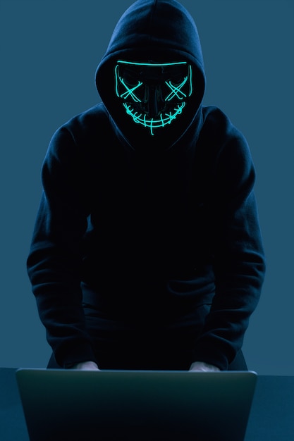 Hombre anónimo con una sudadera negra y una máscara de neón hackeando una computadora