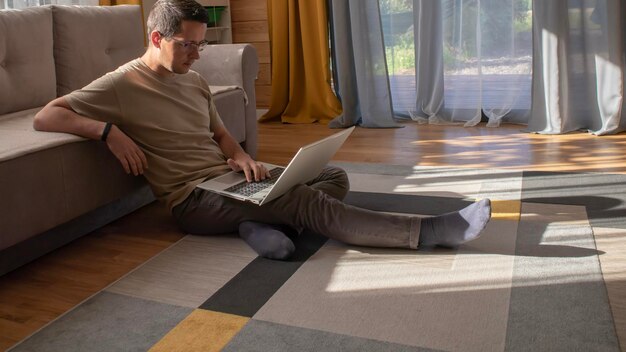 El hombre con anteojos con una computadora portátil está trabajando desde casa sentado en el suelo en un interior moderno y elegante