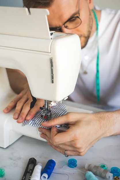 Un hombre con anteojos está cosiendo en una máquina de coser Un sastre con una cinta métrica en el cuello con una camiseta blanca El concepto de estilo de vida de taller en el hogar de pequeñas empresas y disfrutar del trabajo