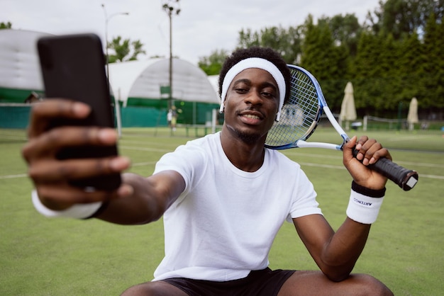 El hombre de apariencia africana está sentado en la hierba de la cancha de tenis sosteniendo el teléfono en un estuche negro sonriendo