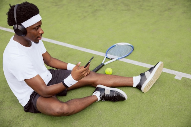 El hombre de apariencia africana está sentado en la hierba de la cancha de tenis sosteniendo el teléfono y usando una diadema