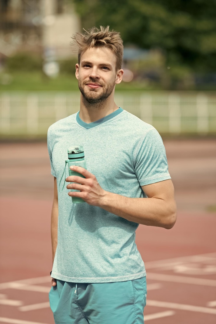 Foto hombre con apariencia atlética sostiene una botella con agua el atleta bebe agua después de entrenar en el estadio en un día soleado deporte y concepto de estilo de vida saludable hombre atleta en ropa deportiva entrenando al aire libre
