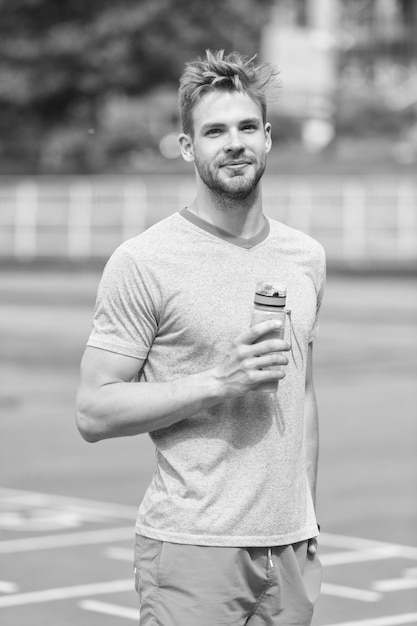 Hombre con apariencia atlética sostiene una botella con agua El atleta bebe agua después de entrenar en el estadio en un día soleado Deporte y concepto de estilo de vida saludable Hombre atleta en ropa deportiva entrenando al aire libre