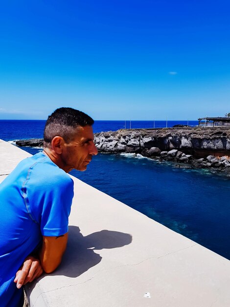 Foto hombre apoyado en la pared de contención mientras mira al mar contra el cielo azul claro