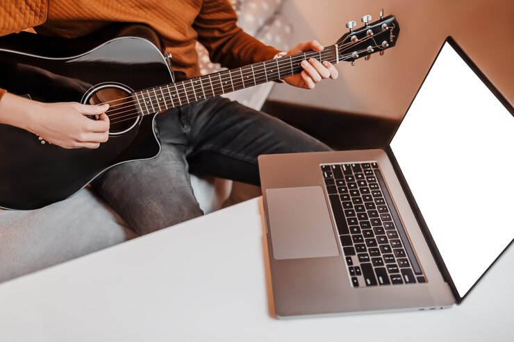 Foto hombre aprendiendo a tocar la guitarra con la ayuda del aprendizaje online en casa. chico sentado a la mesa con laptop y guitarra negra