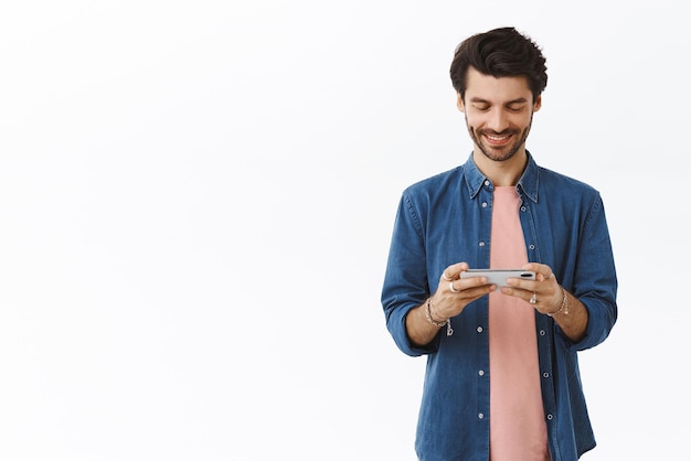 Un hombre apuesto y alegre que se relaja durante la pausa del almuerzo jugando un nuevo y genial juego móvil sosteniendo el teléfono inteligente con ambas manos en posición horizontal sonriendo encantado mientras mira fijamente el fondo blanco de la pantalla del teléfono