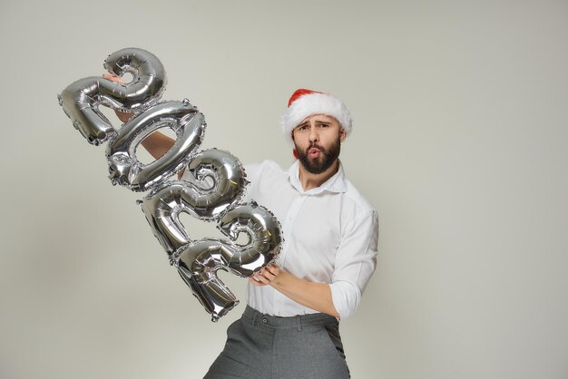 Un hombre apuesto con un gorro de Papá Noel de terciopelo rojo con los labios metidos en un tubo empuja globos plateados con la forma de 2022 hacia la vertical a la izquierda. Un chico con barba en una fiesta de año nuevo.