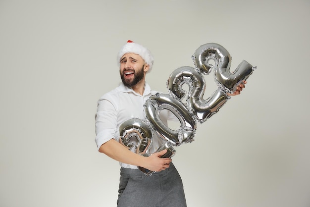 Un hombre apuesto con un gorro de Papá Noel de terciopelo rojo sostiene globos plateados con la forma de 2022 como una ametralladora. Un chico sonriente con barba en una fiesta de año nuevo.