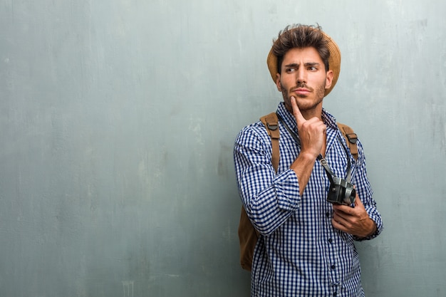Hombre apuesto joven del viajero que lleva un sombrero de paja, una mochila y una cámara de la foto pensando y mirando para arriba