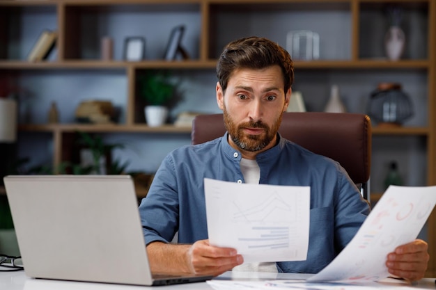 Un hombre apuesto que trabaja en una laptop mientras está sentado en la oficina de su casa Con estrés mira los gráficos de ventas La crisis financiera en la bolsa de valores Malas noticias para los negocios El colapso de la empresa