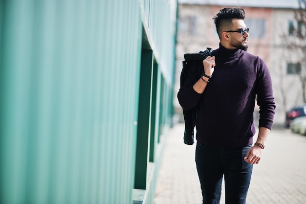 Hombre árabe de barba rica en moda con cuello de tortuga y gafas de sol caminando contra la pared verde con chaqueta en la mano Elegante modelo árabe exitoso y de moda