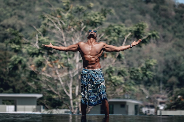 Hombre árabe sin camisa en forma y guapo con pareo turquesa y gafas de sol de pie junto a la piscina en un día soleado, con los brazos abiertos; palmas y cielo; concepto de paraíso.