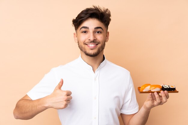 Hombre árabe comiendo sushi aislado en la pared beige con los pulgares hacia arriba porque ha sucedido algo bueno