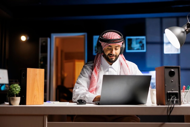 Un hombre árabe confiado escribiendo en una computadora portátil dedicado a comunicaciones, videoconferencias y correo electrónico. Un musulmán escucha y mira con auriculares inalámbricos mientras investiga y navega por Internet.