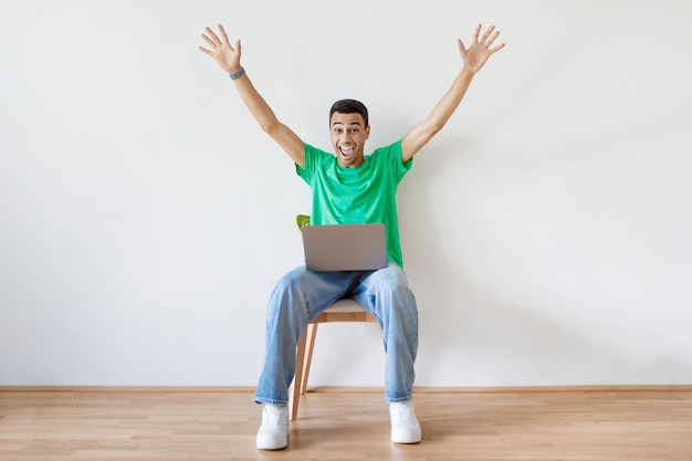 Hombre árabe emocional sentado en una silla con una computadora portátil y levantando las manos, un tipo feliz consiguió el trabajo de sus sueños o una gran noticia