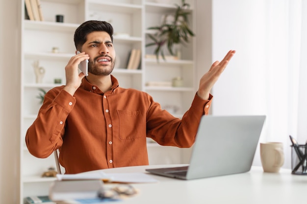 Hombre árabe enojado hablando por teléfono celular usando una computadora portátil