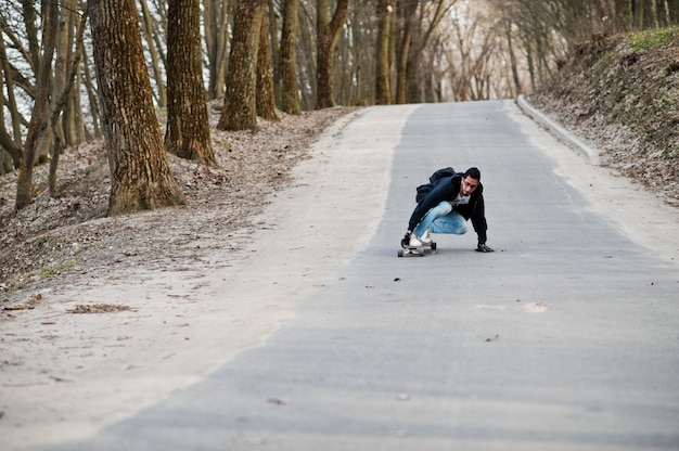 Hombre árabe de estilo callejero en anteojos con longboard longboarding por el camino.