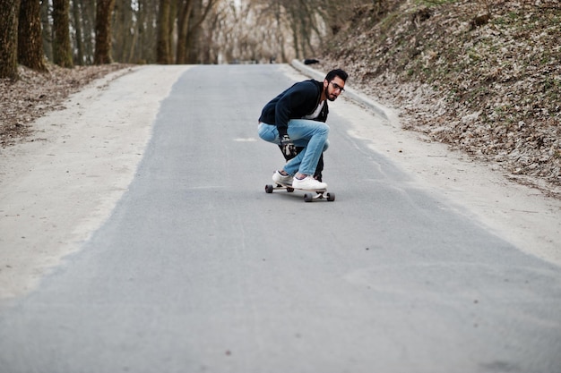 Hombre árabe de estilo callejero en anteojos con longboard longboarding en el camino